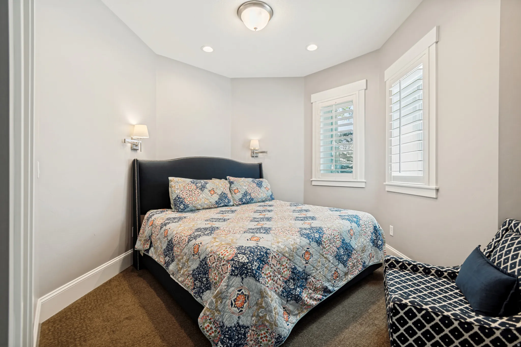Carpeted room featuring dual wall sconces, ceiling-mounted lighting, and two windows with plantation shutters