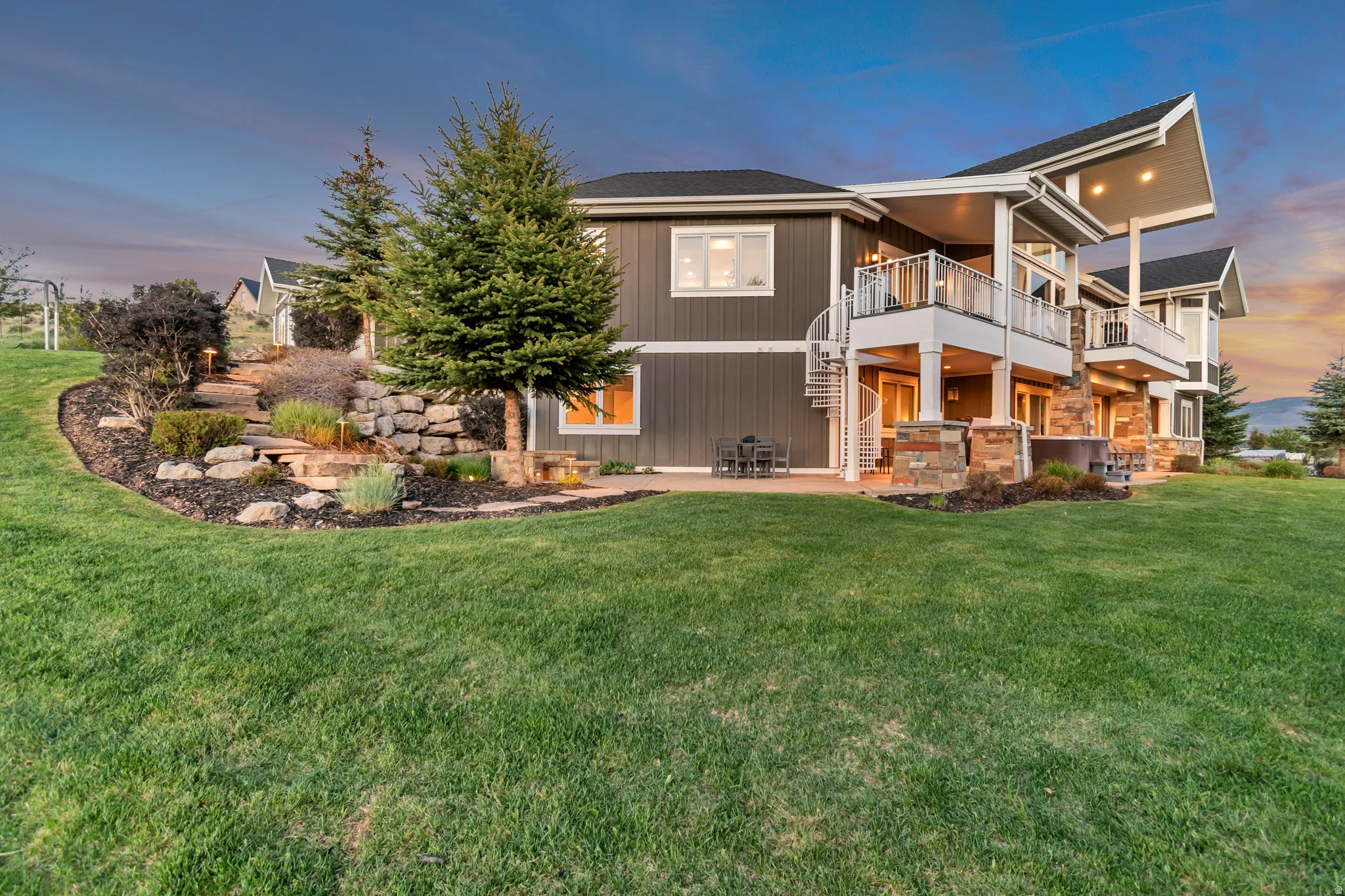 Expansive green lawn surrounds a multi-level residence featuring a covered patio, a spiral staircase, and an upper-level balcony with glass railings