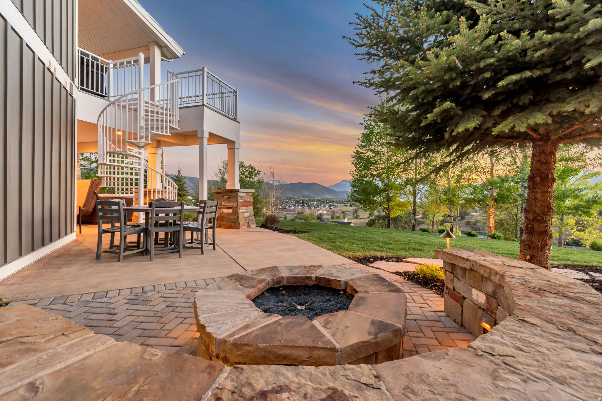 Expansive outdoor patio featuring a built-in fire pit with stone surround, a curved stone bench, and a spiral staircase to an upper-level balcony
