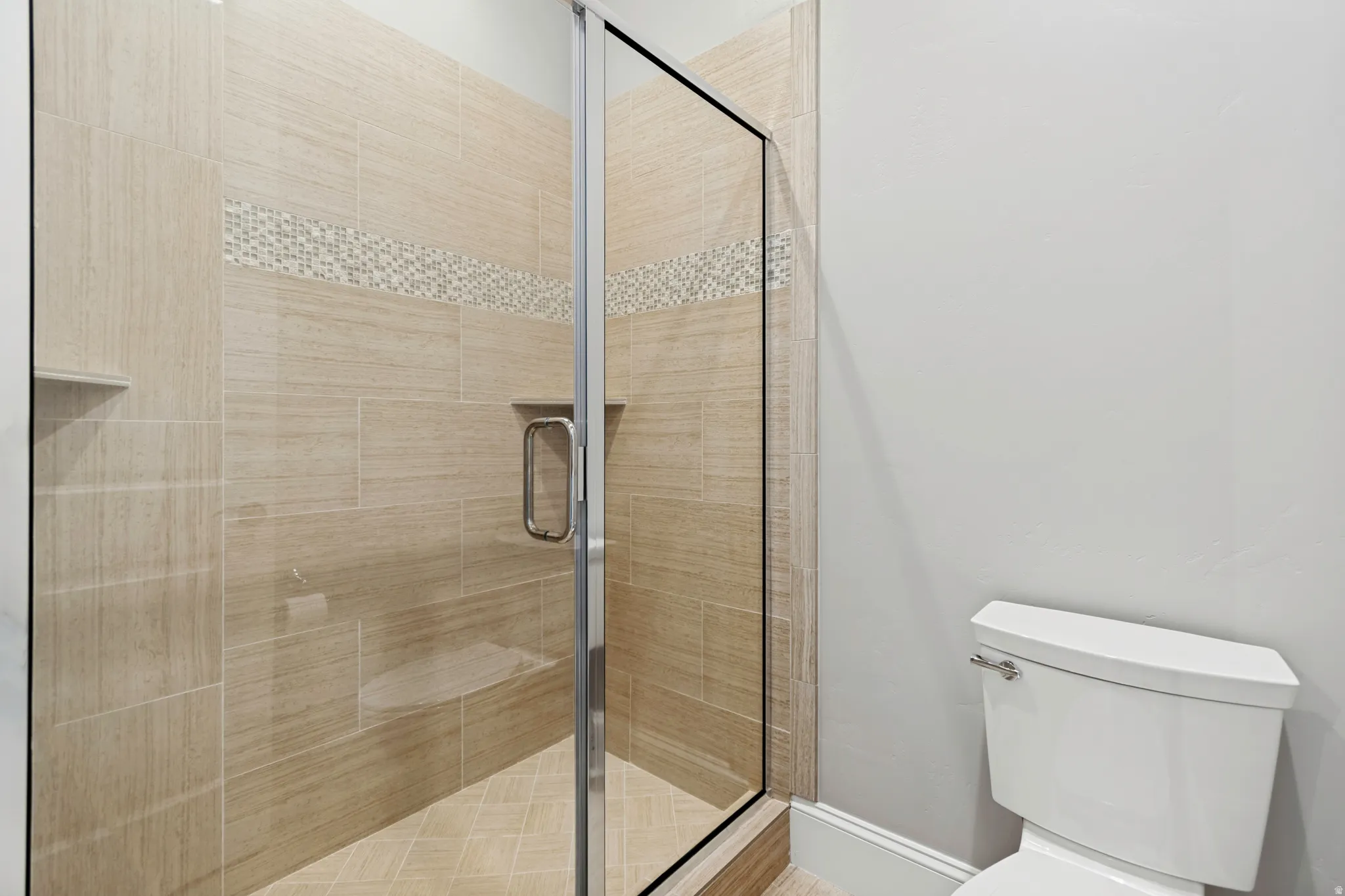 Walk-in shower featuring glass enclosure, light-toned tile walls with a decorative mosaic accent, a built-in shower bench, and recessed shelving