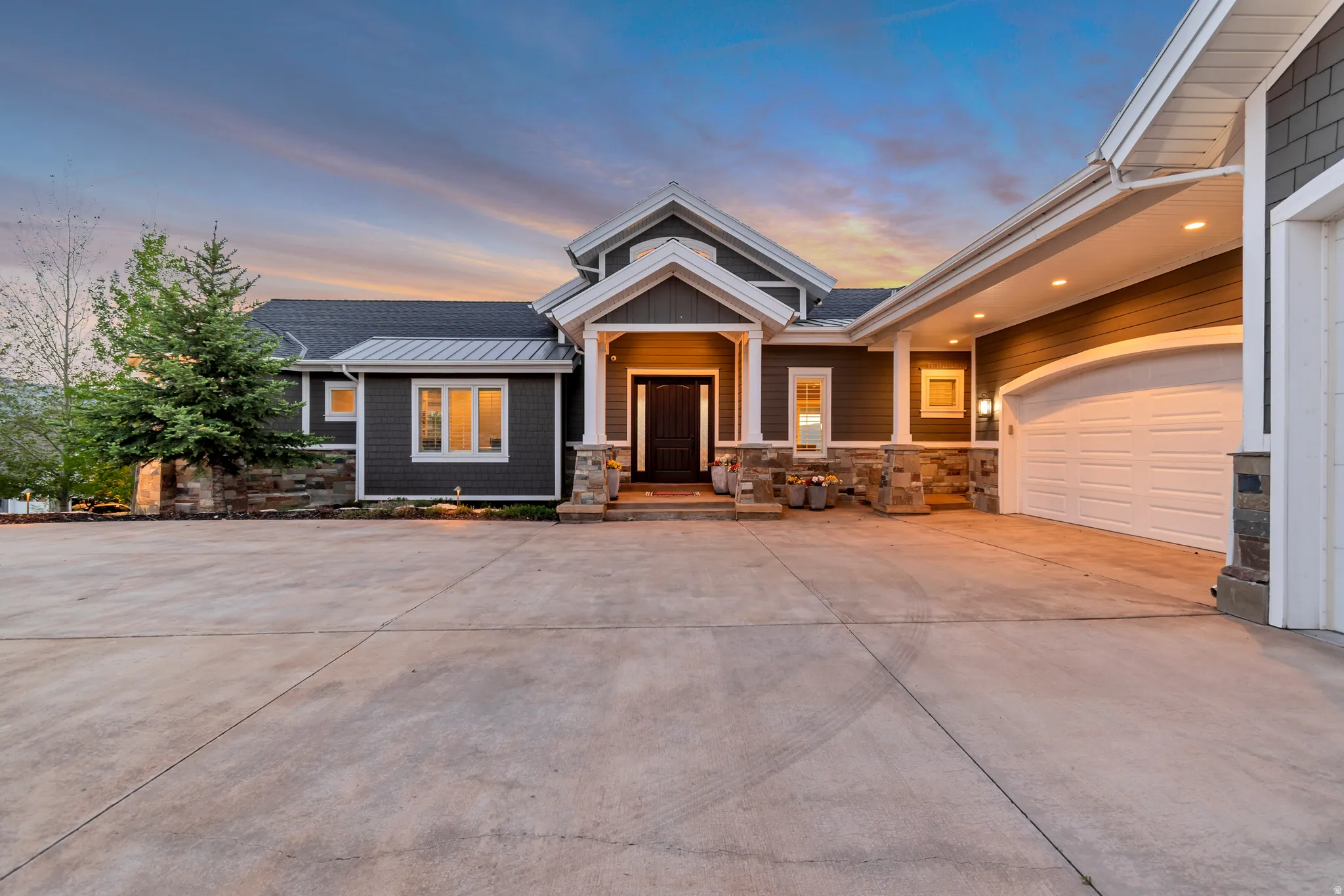 Contemporary exterior featuring dark gray siding, stone accents, a prominent front entry with an overhang, a multi-car garage with arched detailing, and a spacious concrete driveway