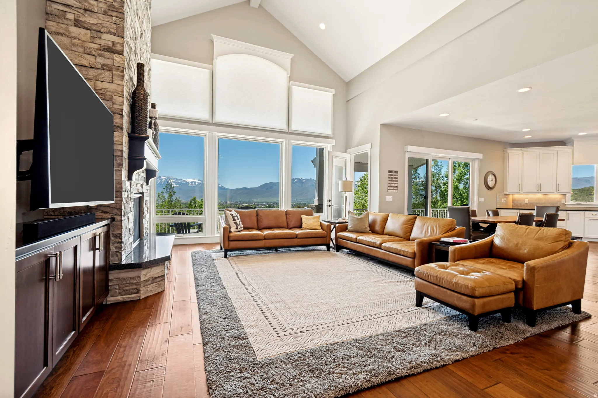 Vaulted ceilings with large windows showcasing mountain scenery, a stone-clad fireplace, and wood-finish flooring