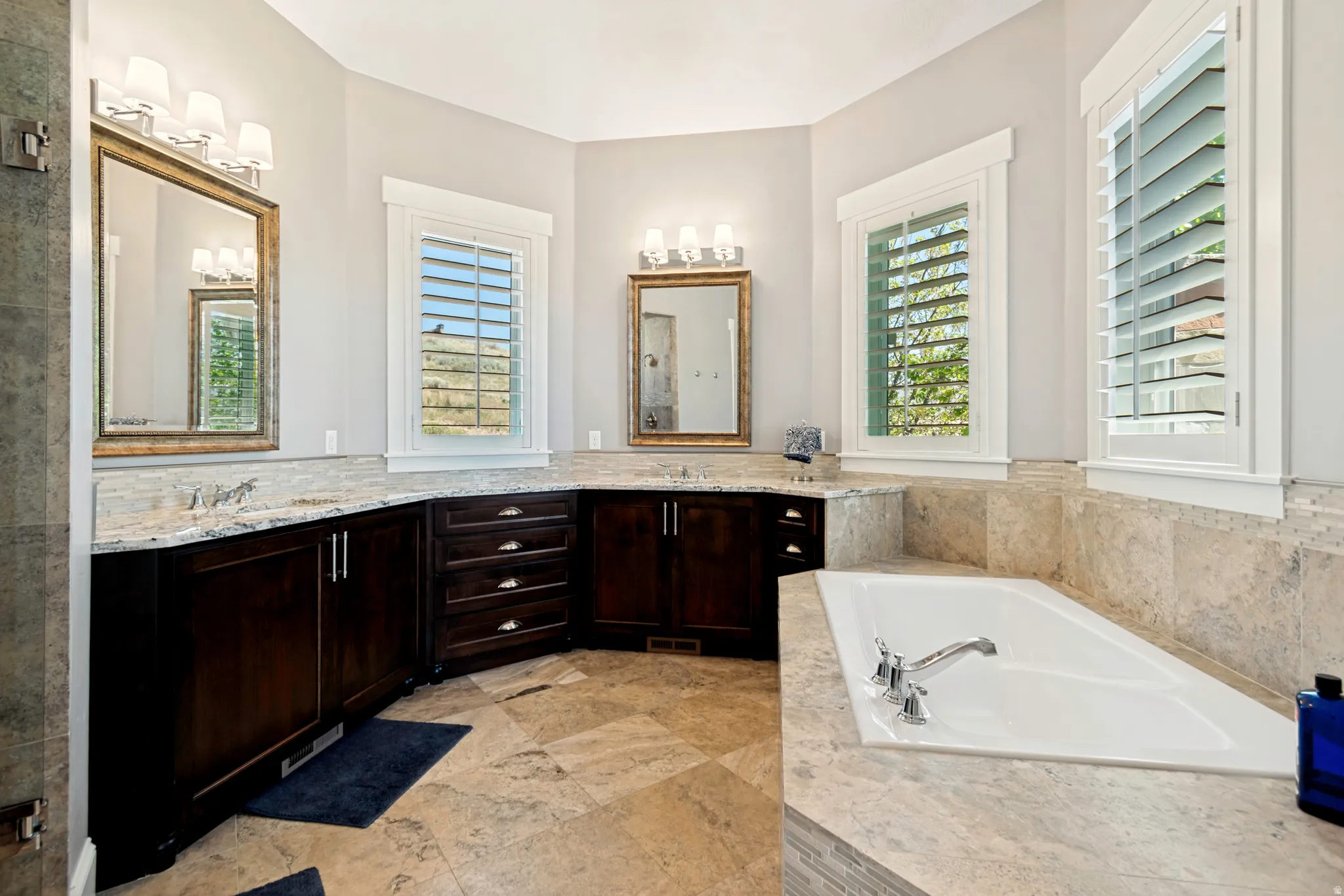 Spacious bathroom featuring a built-in soaking tub, dual vanity areas with dark wood cabinetry, granite countertops, and stone tile flooring