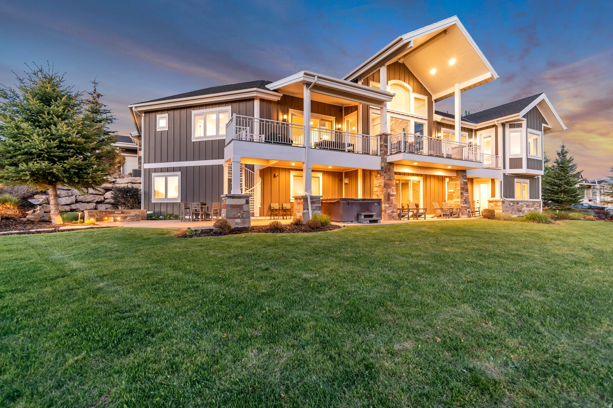 Expansive rear exterior featuring a multi-level design, stone accents, multiple balconies with railings, a walk-out patio with a built-in hot tub, and a spacious lawn