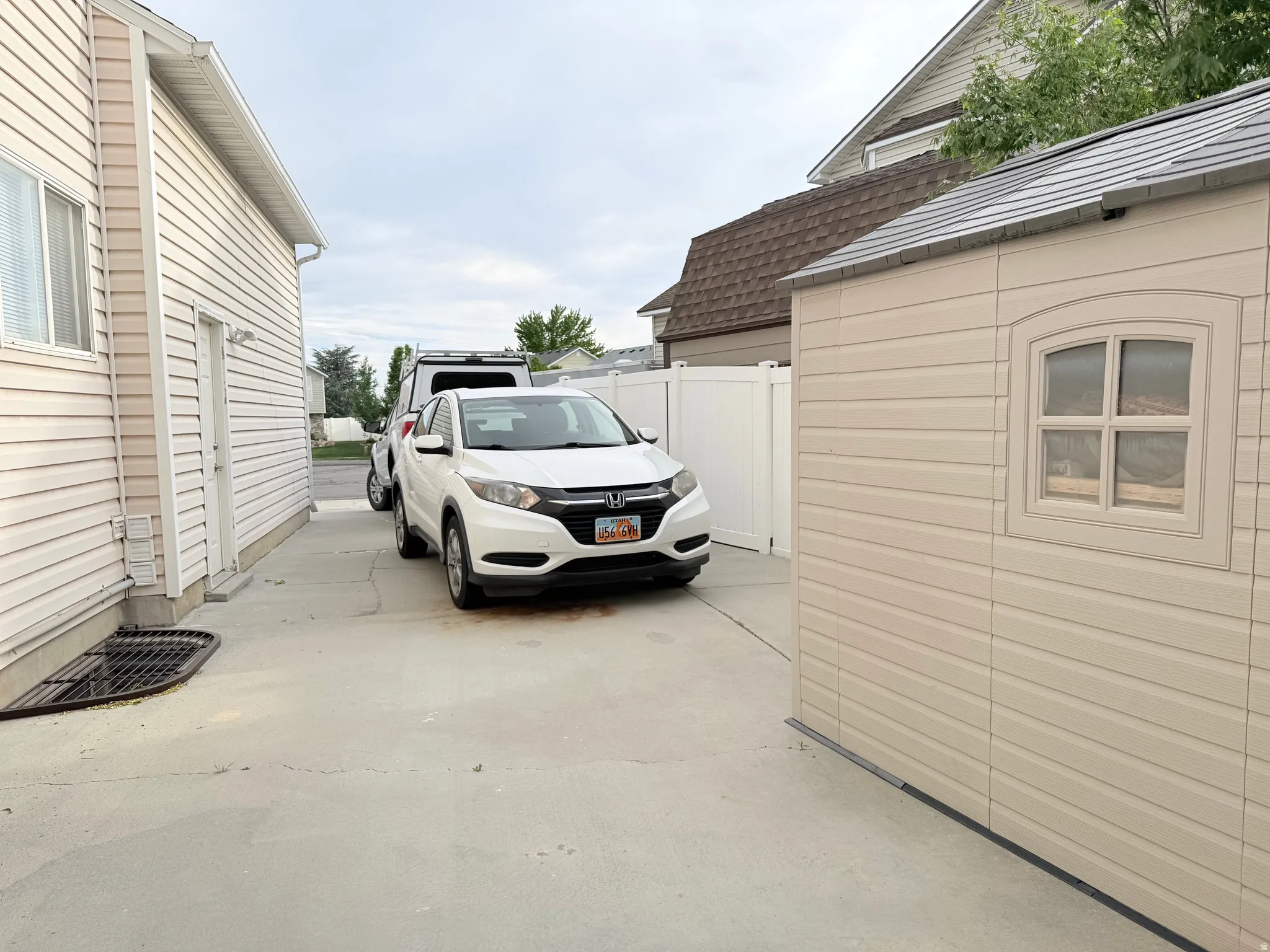 Concrete driveway alongside a beige vinyl-sided structure, featuring a white privacy fence and a shed with an arched window