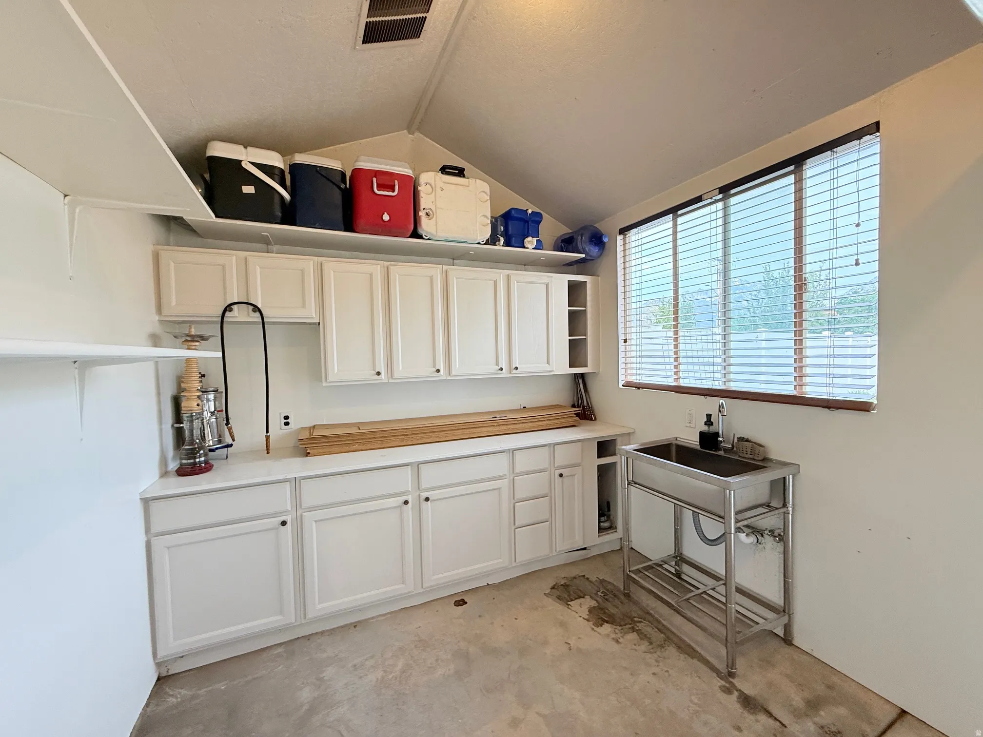 Utility room featuring extensive white cabinetry, a built-in stainless steel sink with a workstation, a large window with horizontal blinds, and a vaulted ceiling with an overhead storage shelf