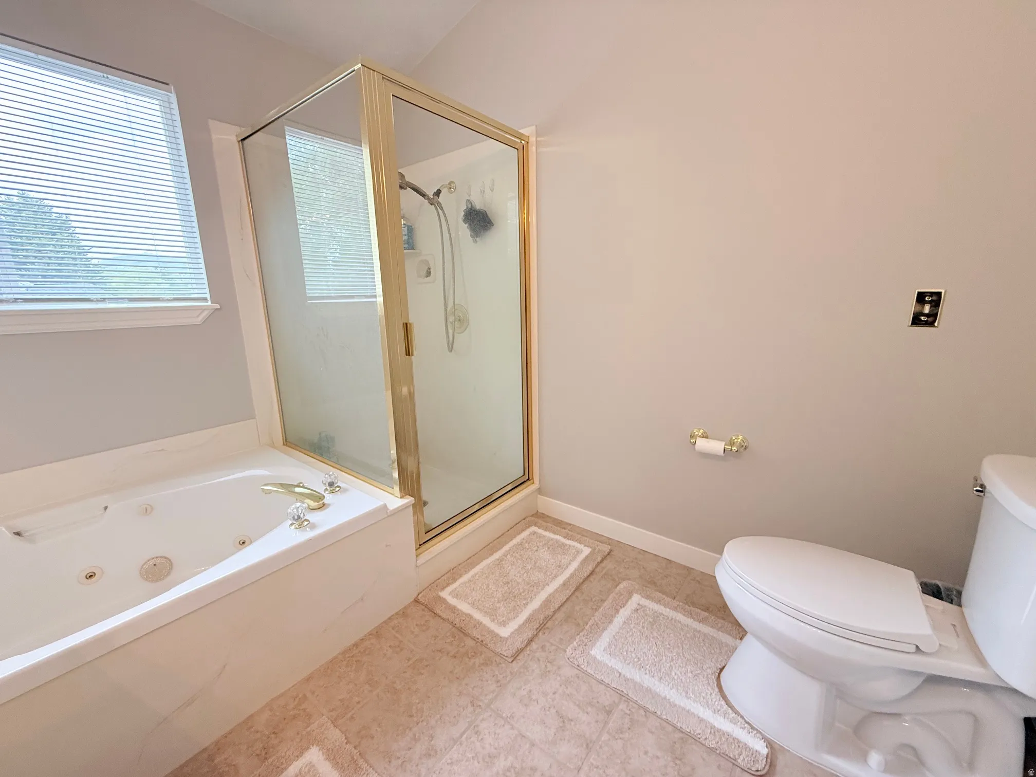 Bathroom featuring a jetted tub, a glass-enclosed shower with brass-finish framing, a window with blinds, and neutral-toned wall paint