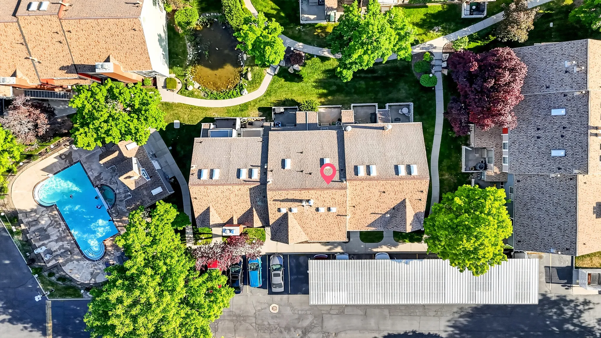 Aerial perspective showcasing a community pool, lush green spaces with mature trees, a tranquil pond feature, and covered parking facilities