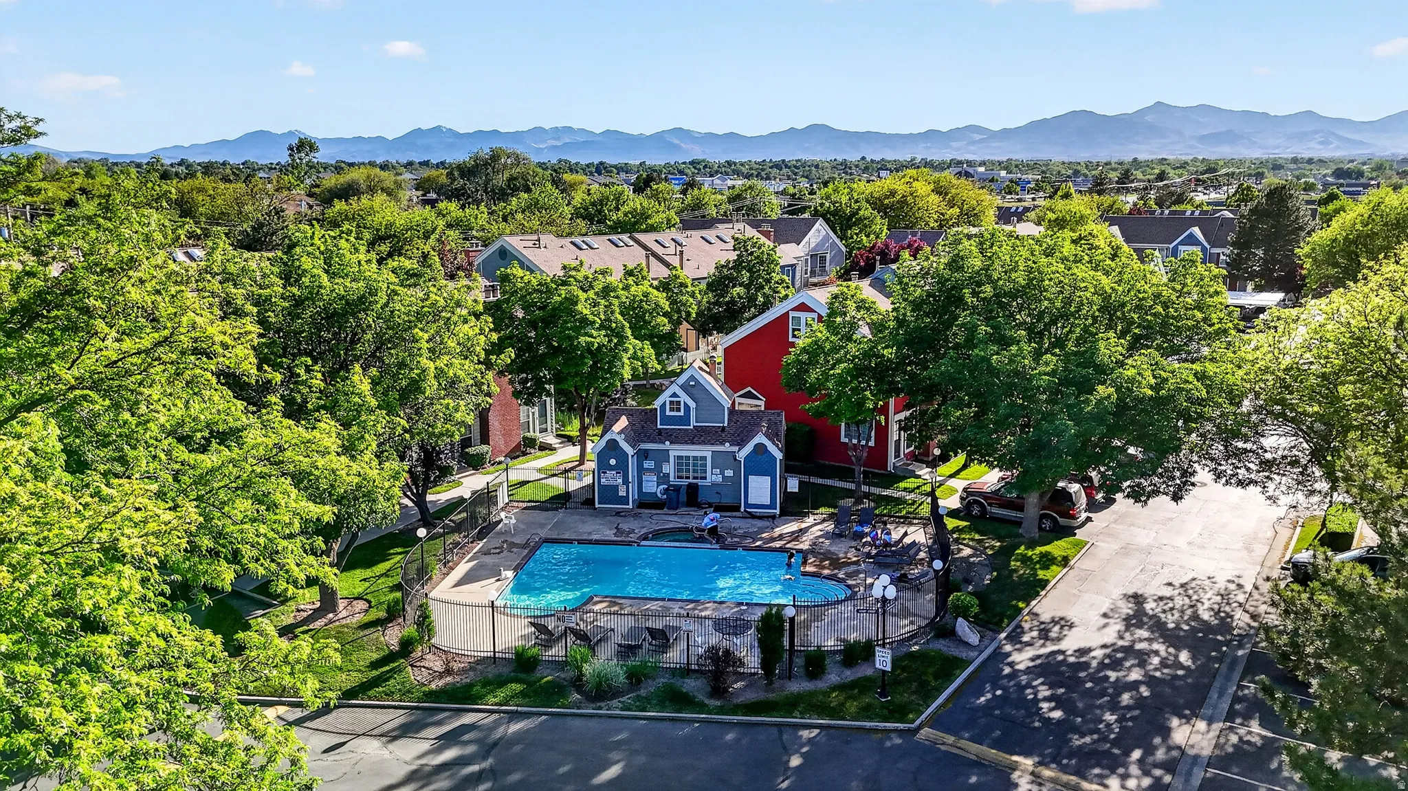 Community swimming pool with surrounding fence and paved patio area, featuring a blue accessory building with white trim