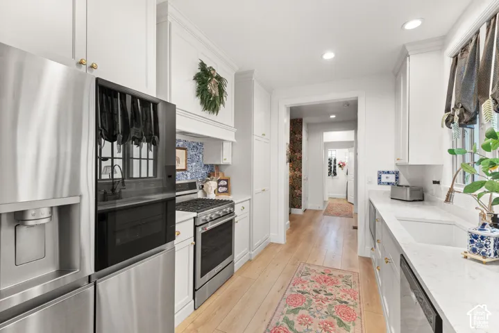 Galley kitchen featuring stainless steel appliances, light wood-finish flooring, white cabinetry with gold-toned hardware imported from the UK, white quartz countertops, and recessed lighting