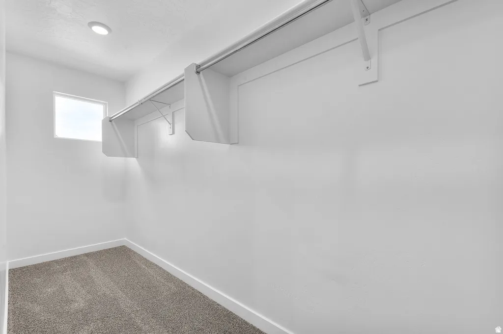 Recessed ceiling lighting illuminating a walk-in closet with a small window, built-in shelving, hanging rod, and gray carpet flooring