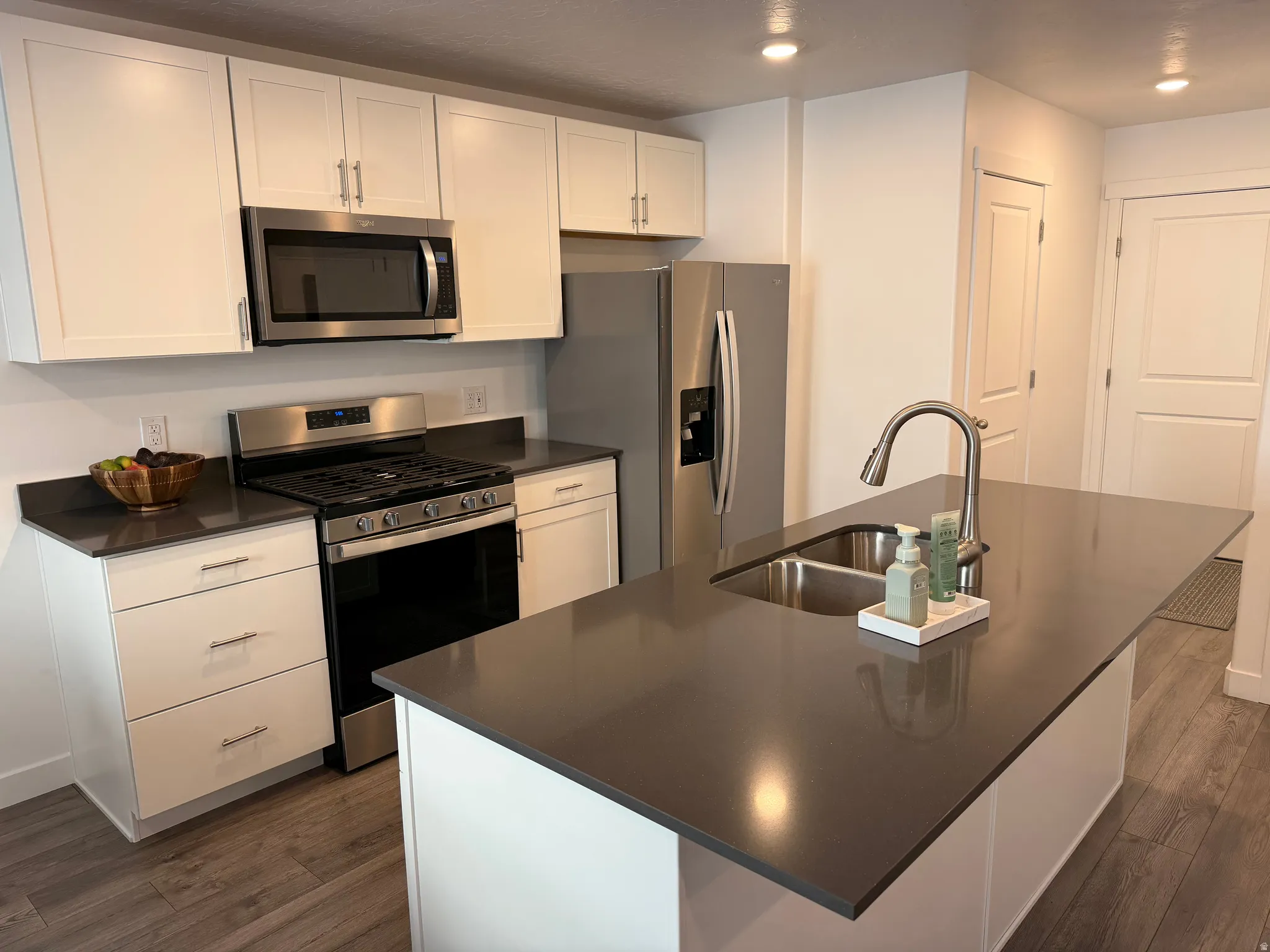 Kitchen featuring an island with an undermount sink, stainless steel appliances, white cabinetry, dark countertops, and wood-finish flooring