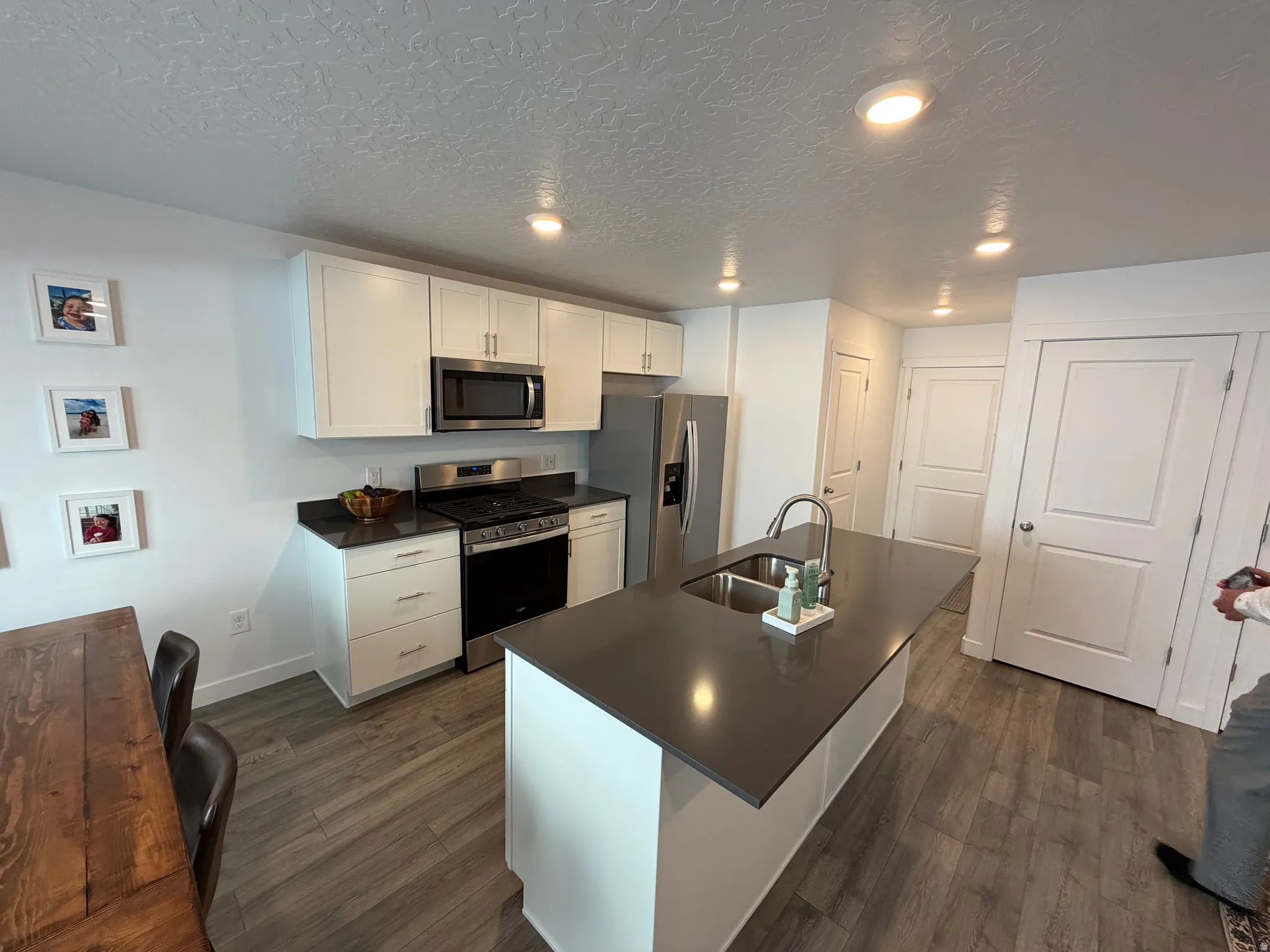 Contemporary kitchen featuring an island with an undermount sink, stainless steel appliances, white cabinetry, dark countertops, and wood-finish flooring