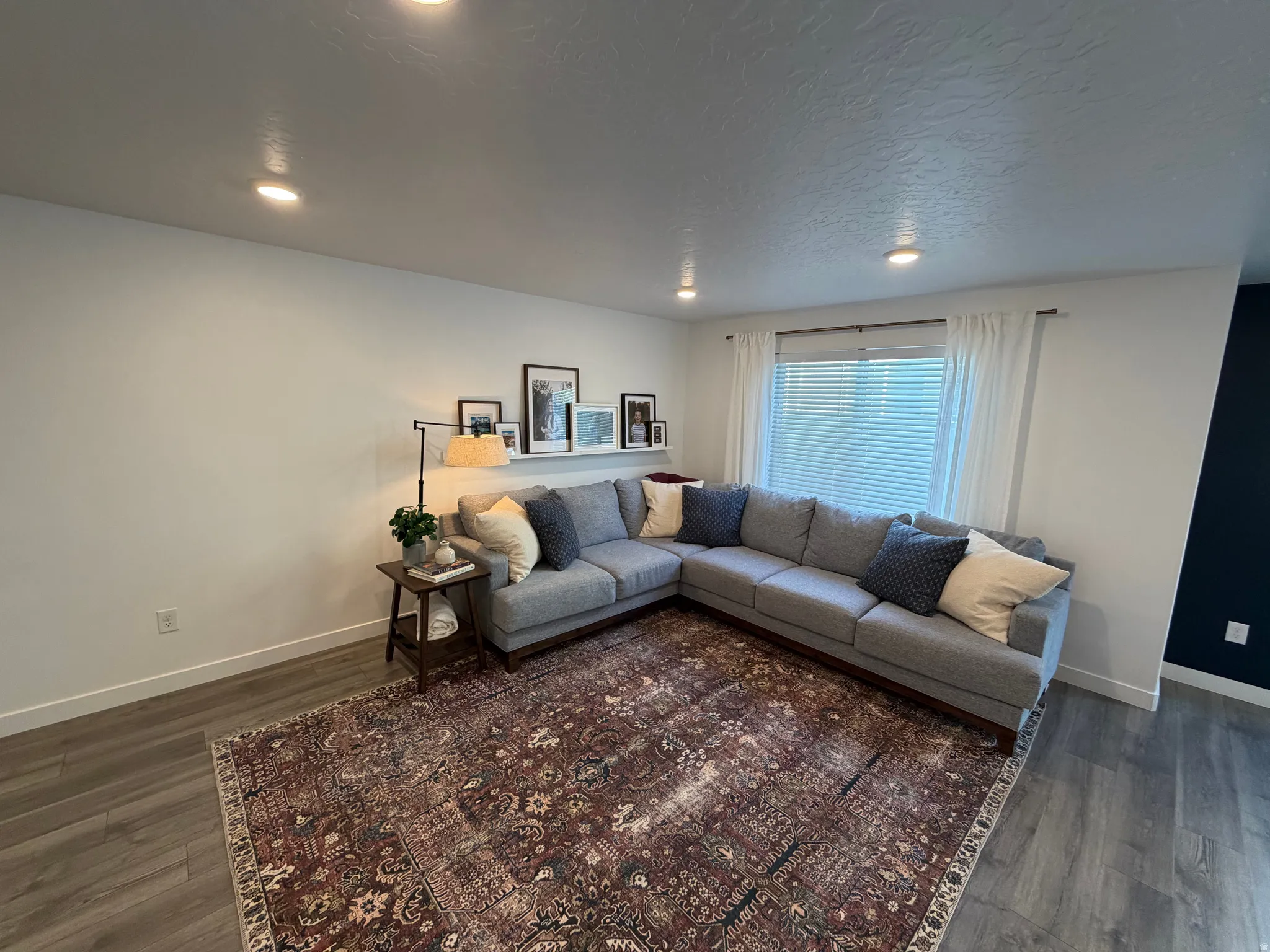 Spacious living area featuring recessed lighting, wood-finish flooring, a large window with blinds and white curtains, and a prominent dark accent wall