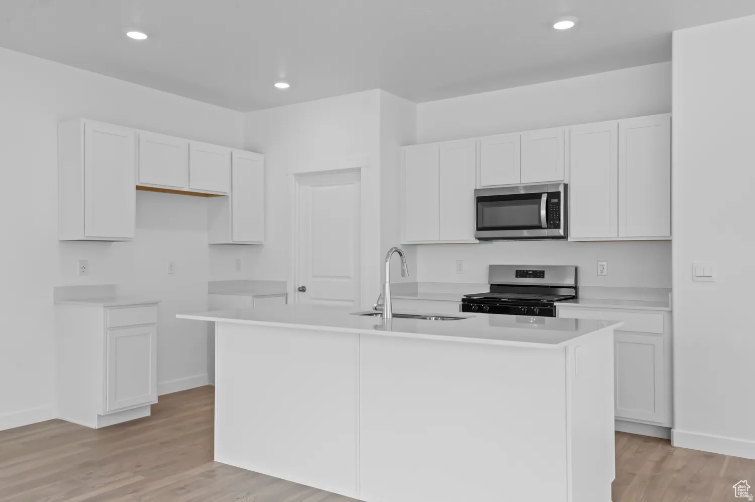 Kitchen featuring white shaker-style cabinetry, stainless steel appliances, a center island with sink, light-toned countertops, and wood-finish flooring