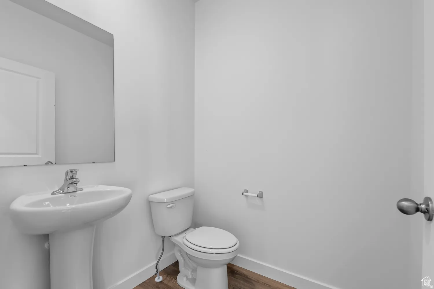 Pedestal sink with a chrome faucet, wall-mounted mirror, toilet, wood-finish flooring, and a brushed nickel door handle