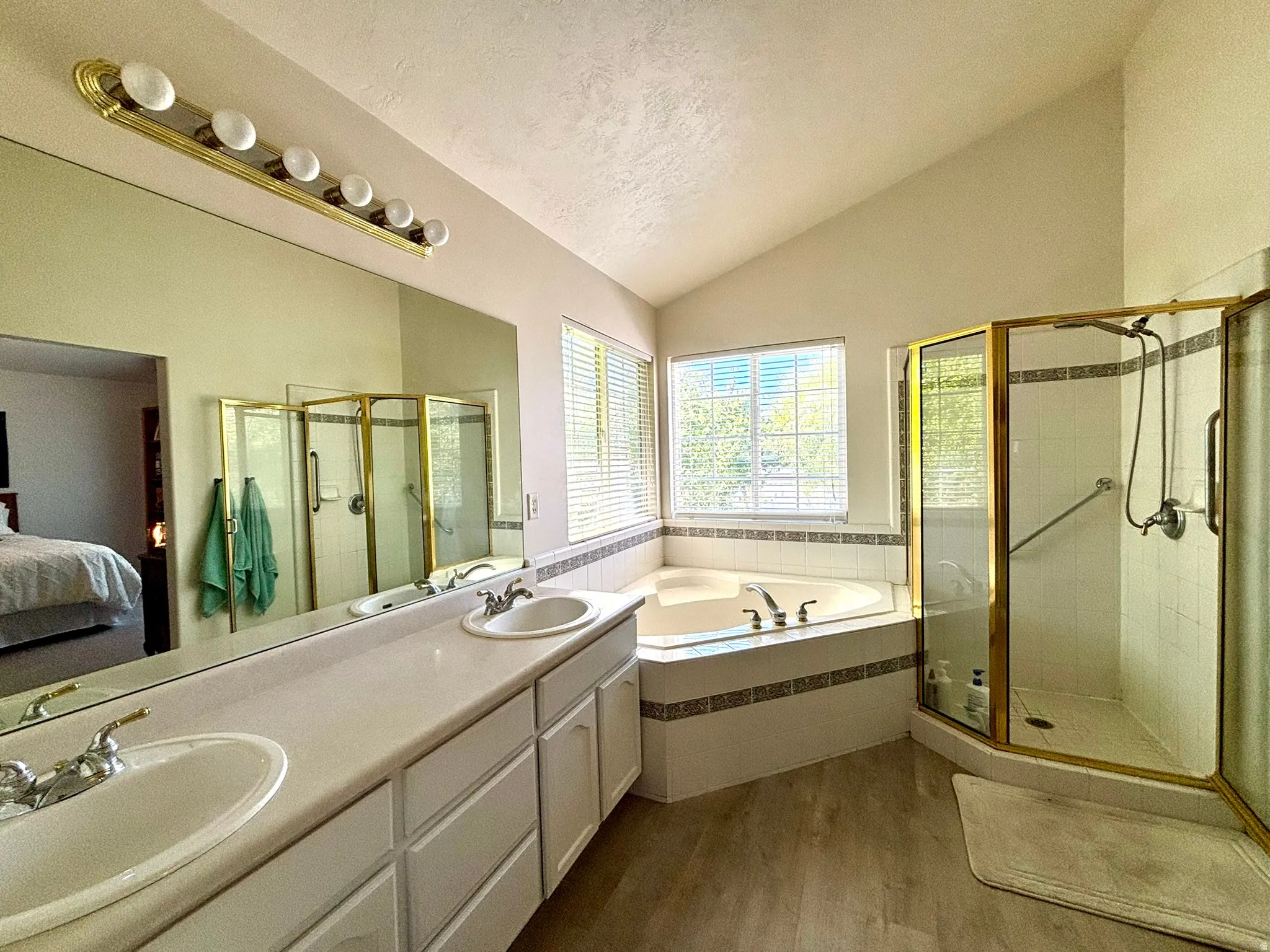 Bathroom featuring dual vanities, large mirror, corner soaking tub, corner shower with gold-tone frame, and vaulted ceiling