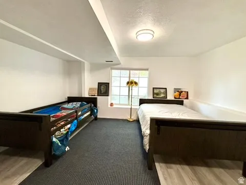 Room featuring dual dark wood-finish bed frames, a window with natural light, a ceiling-mounted light fixture, and a combination of grey carpet and wood-finish flooring
