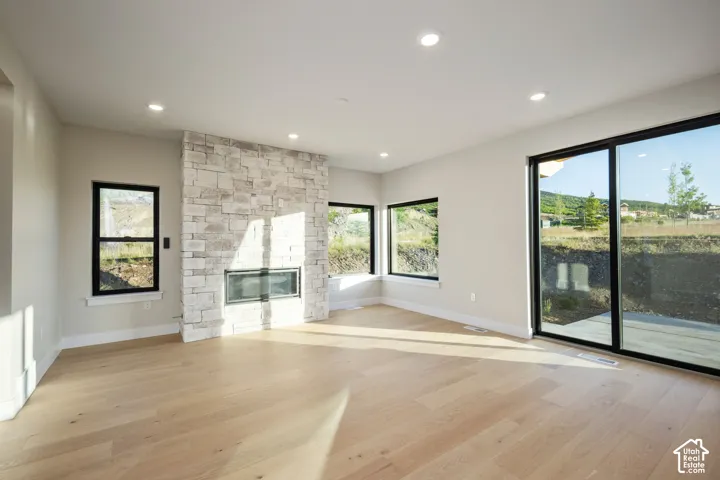 Spacious room featuring light wood-finish flooring, a stone-clad fireplace with a dark-framed insert, multiple large windows, a sliding glass door, and recessed ceiling lighting