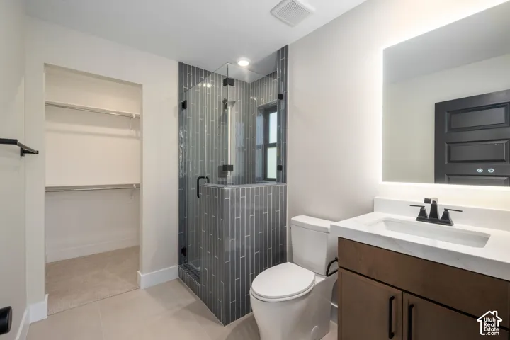 Bathroom featuring a glass-enclosed shower with vertical gray tile, a wood-finish vanity with a white countertop and integrated sink, a backlit mirror, and a walk-in closet with shelving and hanging rods