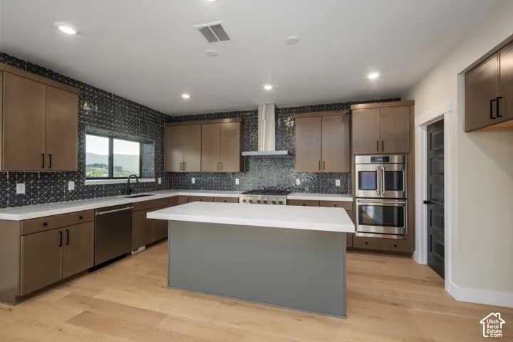 Modern kitchen featuring an island with a white solid surface countertop, light wood-finish flooring, brown cabinetry, stainless steel appliances, and a dark geometric tile backsplash