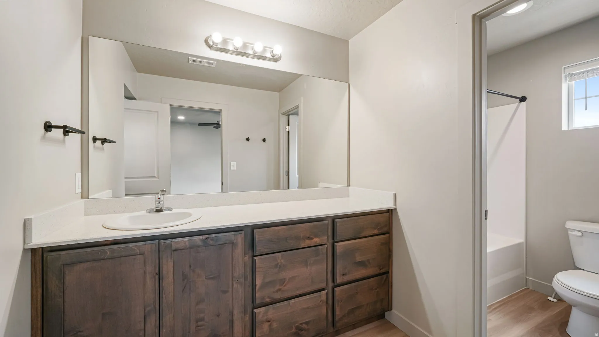 Bathroom featuring a long vanity with a light-colored countertop and integrated sink, rich wood-finish cabinetry, a large wall-mounted mirror, and a contemporary four-bulb light fixture