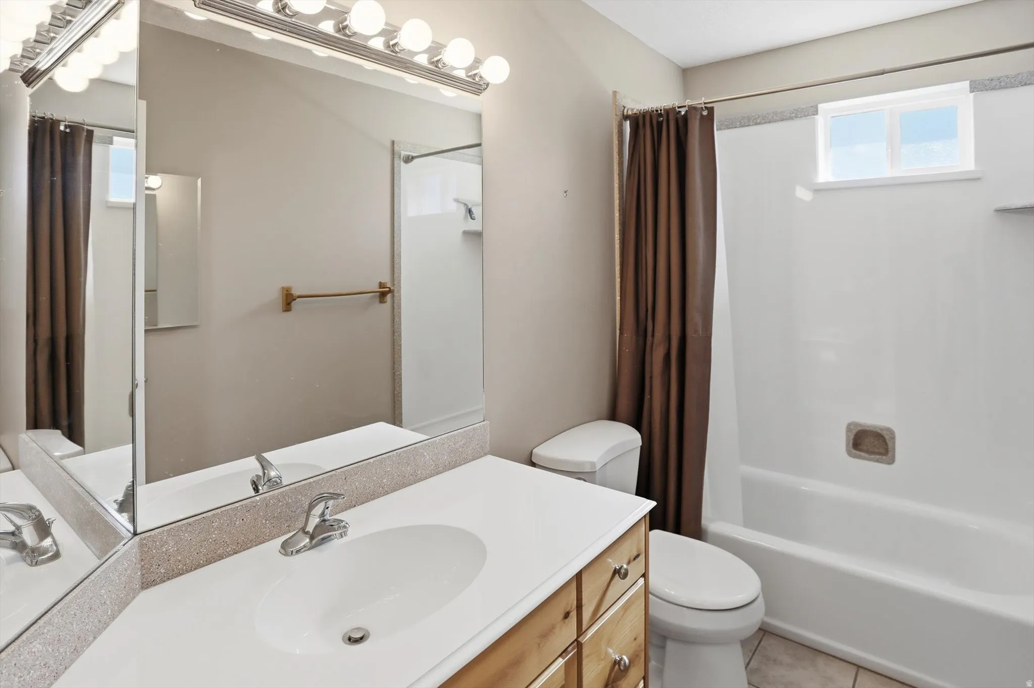 Bathroom featuring a vanity with an integrated sink, a large mirror with a Hollywood-style light fixture, a toilet, and a bathtub/shower combination with an opaque window