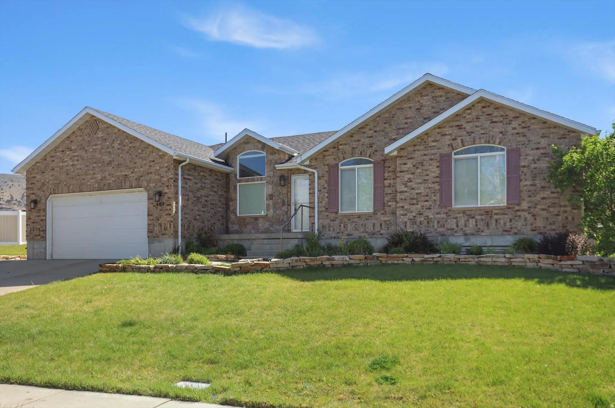 Brick exterior residence featuring a two-car garage, arched windows with shutters, and a stone-edged garden bed