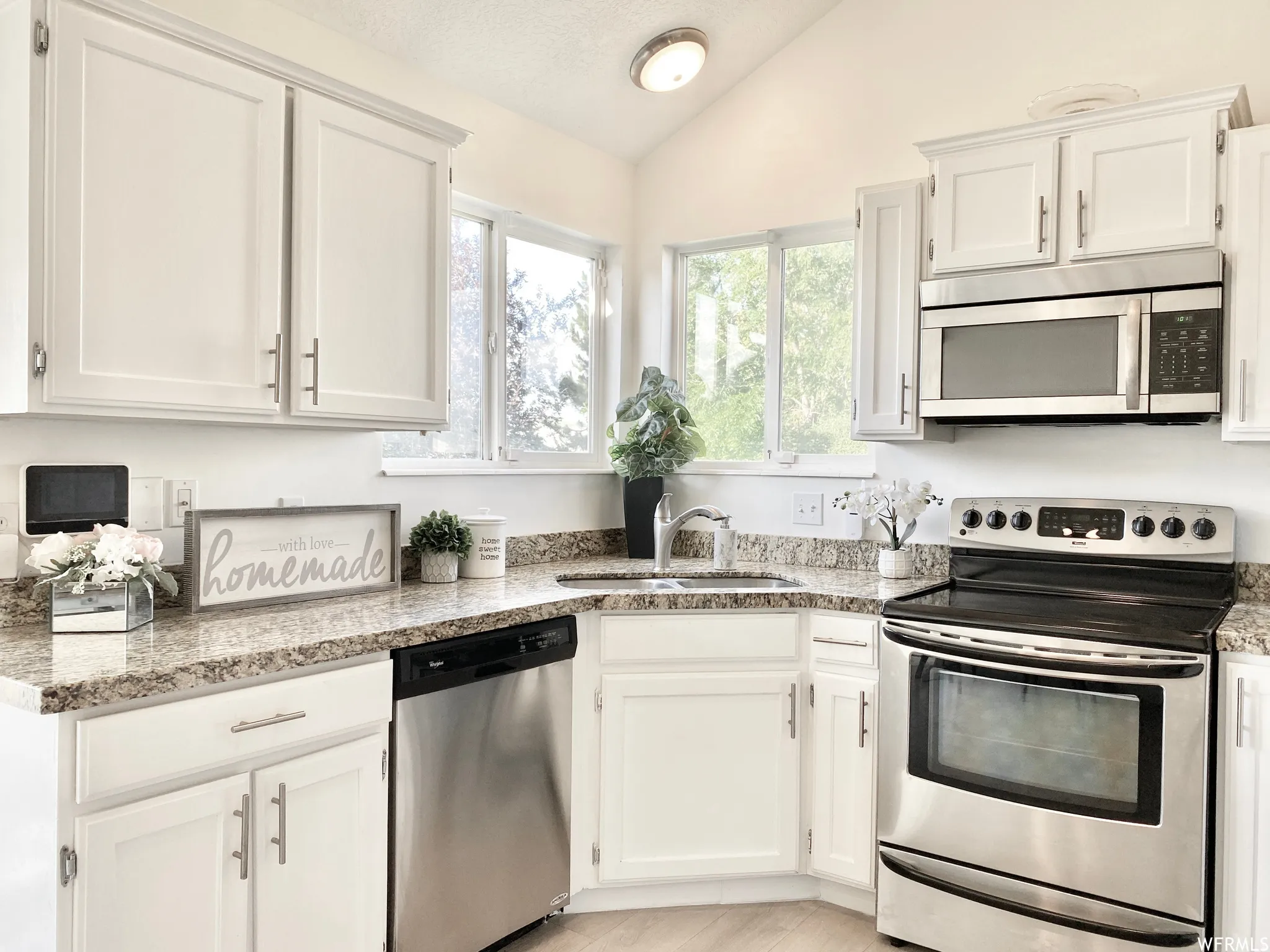 Kitchen featuring granite-style countertops, stainless steel appliances, white cabinetry, a double-basin sink with a gooseneck faucet, and natural light from the corner windows