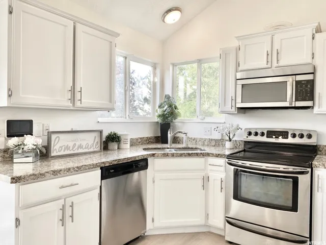 Kitchen featuring granite-style countertops, stainless steel appliances, white cabinetry, a double-basin sink with a gooseneck faucet, and natural light from the corner windows