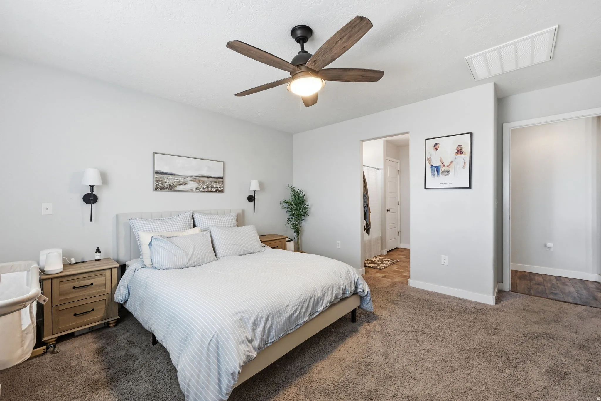 Bright bedroom featuring a ceiling fan with light fixture, dual wall sconces, plush carpet flooring, and a recessed ceiling vent
