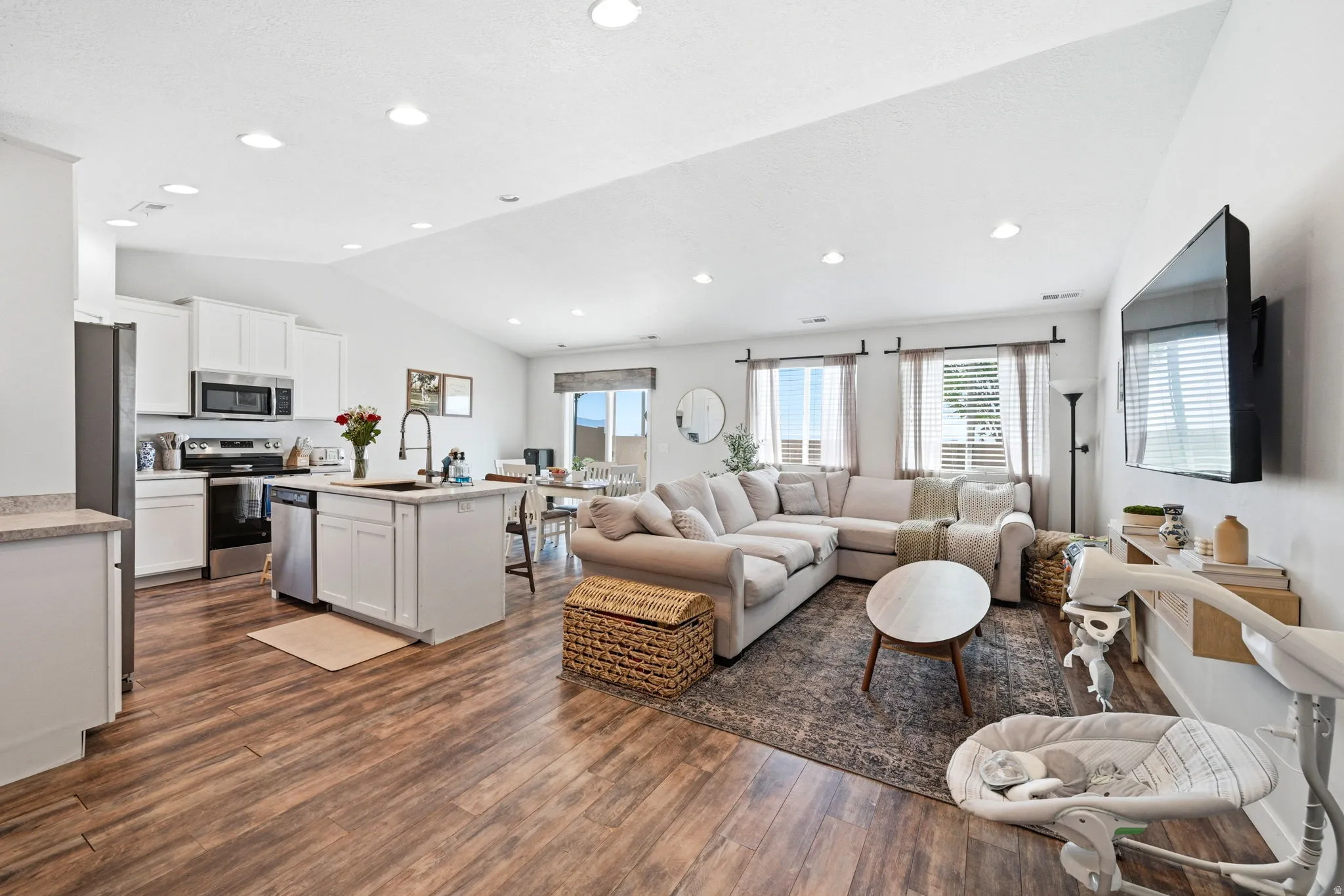 Open concept living space featuring wood-finish flooring, vaulted ceiling, recessed lighting, and a kitchen with white cabinetry, stainless steel appliances, and an island with an undermount sink