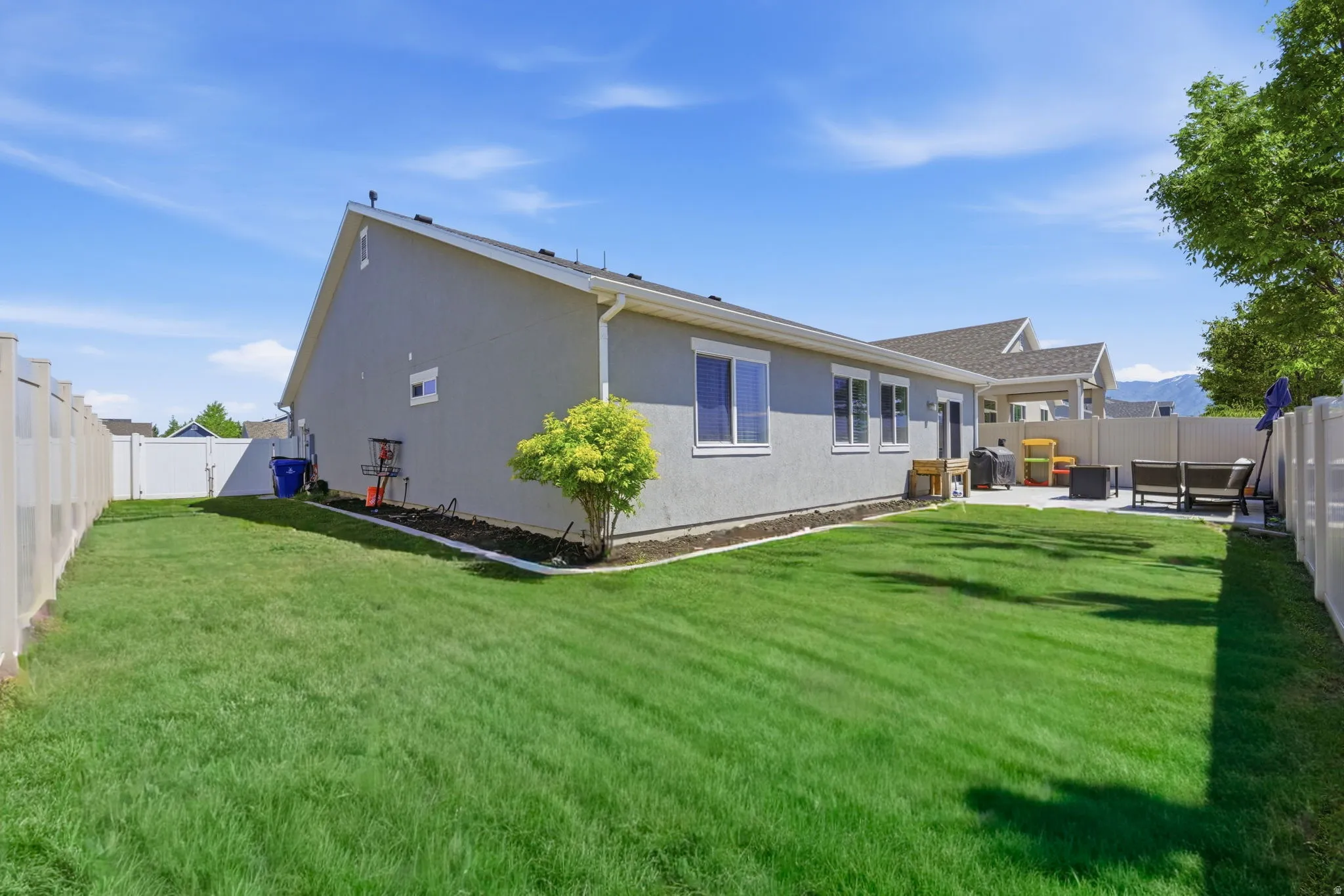 Spacious rear yard with a neutral-toned stucco exterior, white vinyl fencing, a concrete patio, and a mature tree providing shade