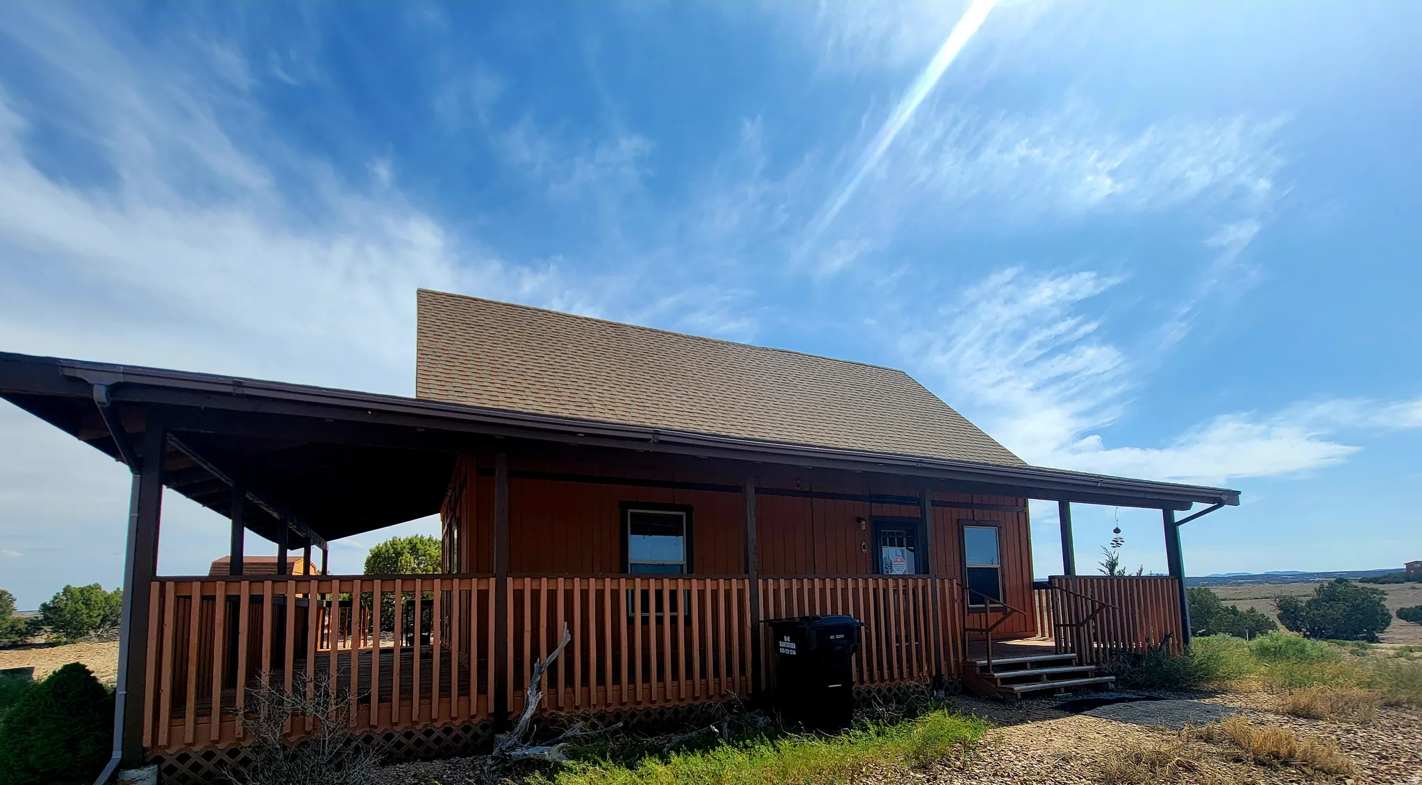 Wood-sided residence featuring an expansive covered porch with wood railing, a brown shingled roof, and a visible desert landscape