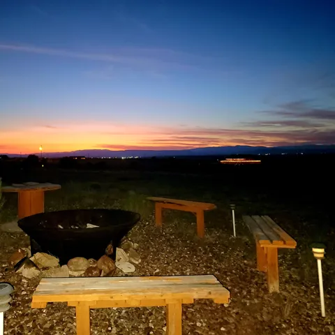 Outdoor fire pit area featuring a large, dark metal fire bowl surrounded by natural stones, three rustic wooden benches, and gravel ground cover