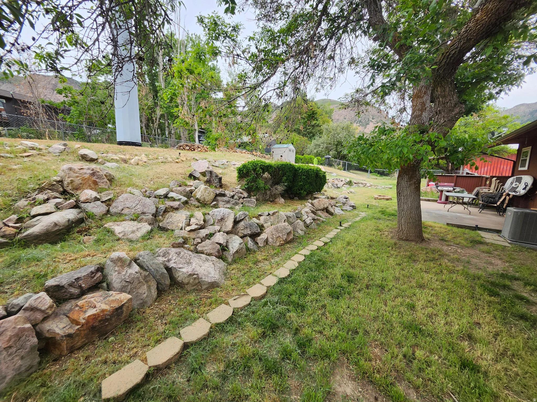 Expansive outdoor space featuring a rock wall, a mature tree, a paved patio, and a grass lawn