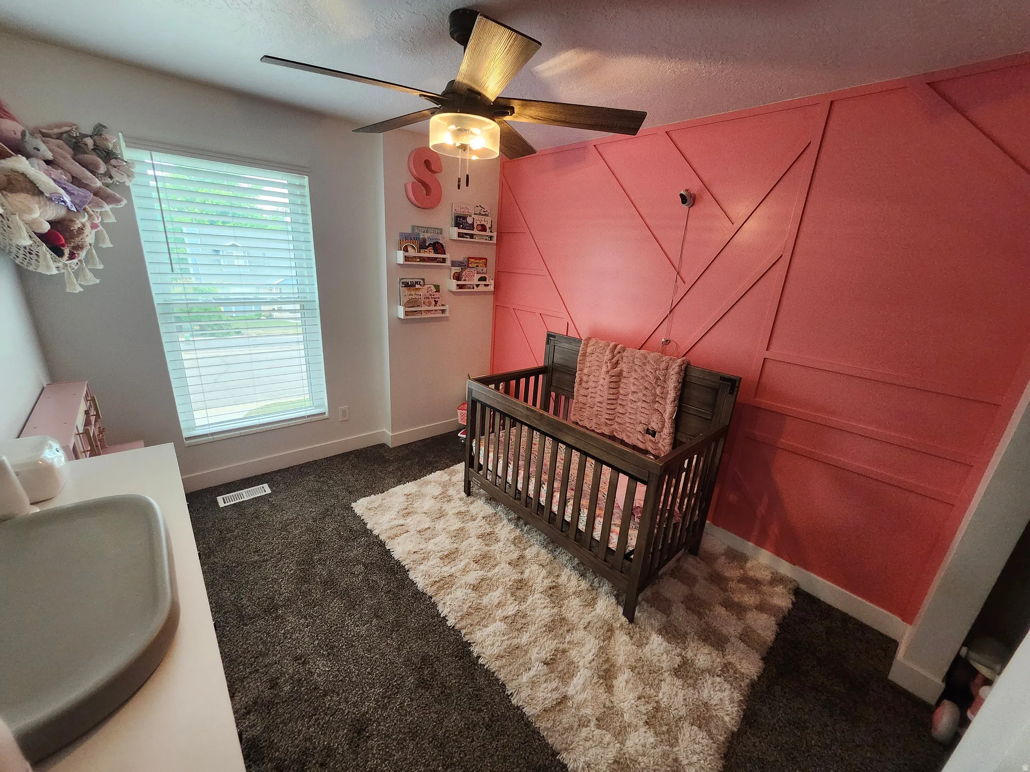 Room featuring a decorative accent wall with geometric wood trim, a window with blinds, dark gray carpeting, a ceiling fan with integrated lighting, and white baseboards