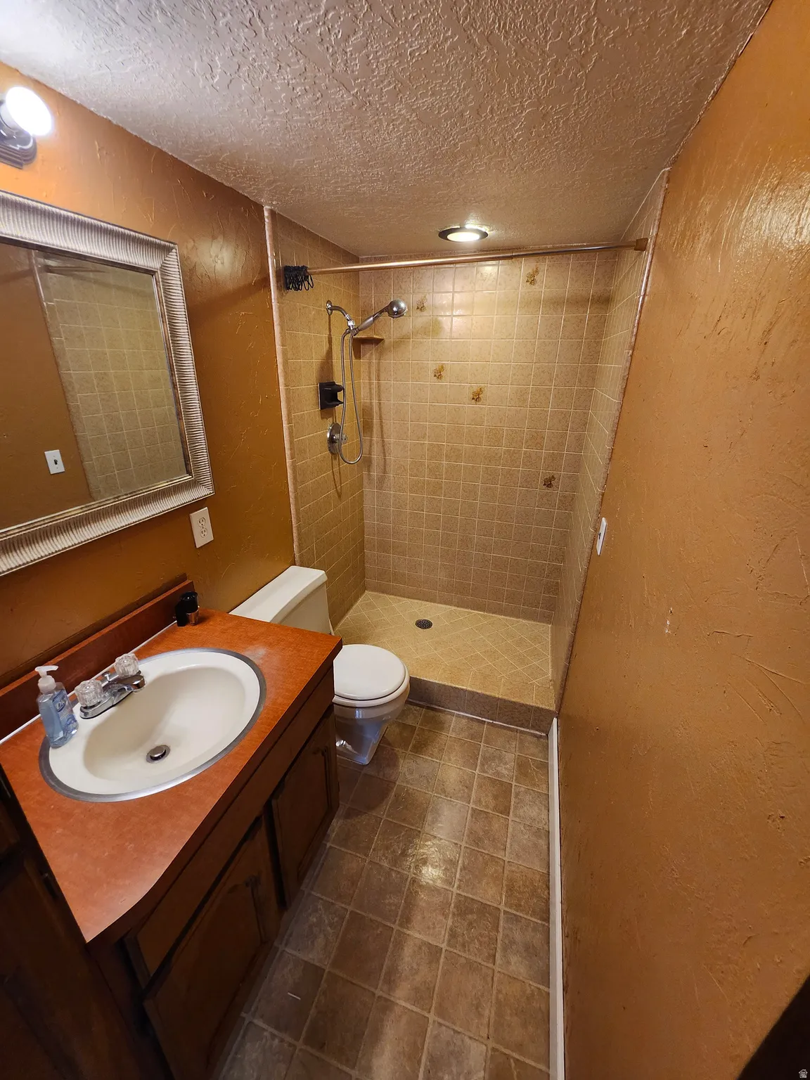 Bathroom featuring a walk-in shower with tiled walls and floor, a single vanity with a white basin and rust-toned countertop, and square floor tiles
