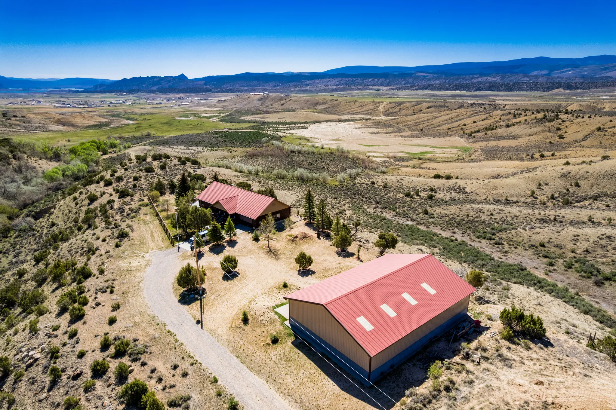 Expansive property featuring two structures with red metal roofs, a winding driveway, and surrounding natural terrain