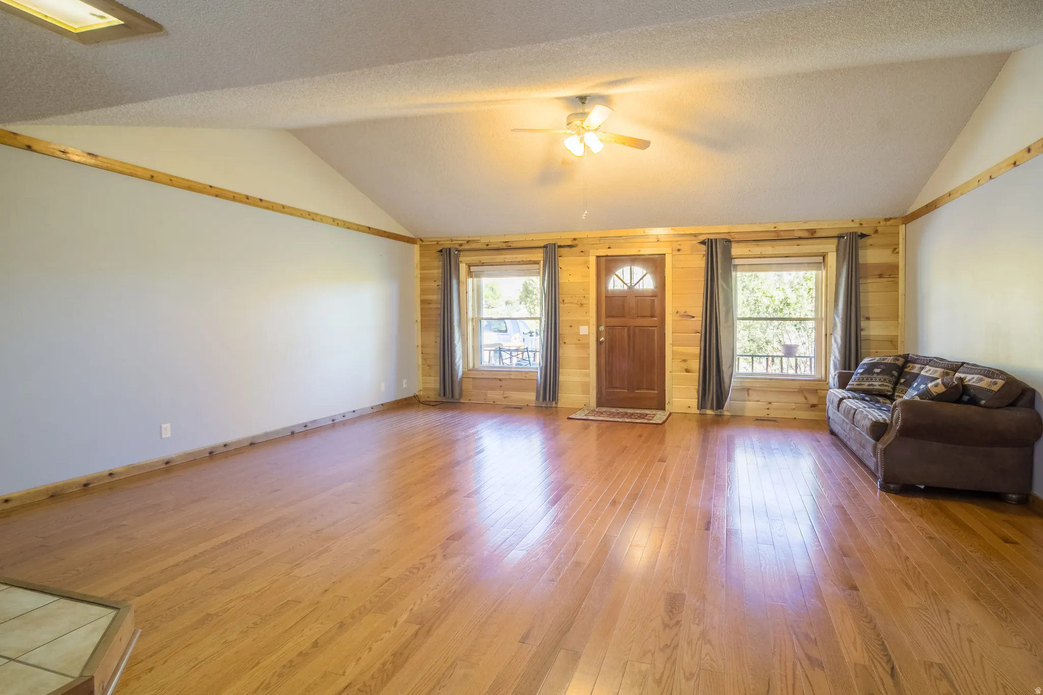 Spacious living area featuring vaulted ceilings, wood-finish flooring, accent wood paneling, a ceiling fan, and abundant natural light from multiple windows