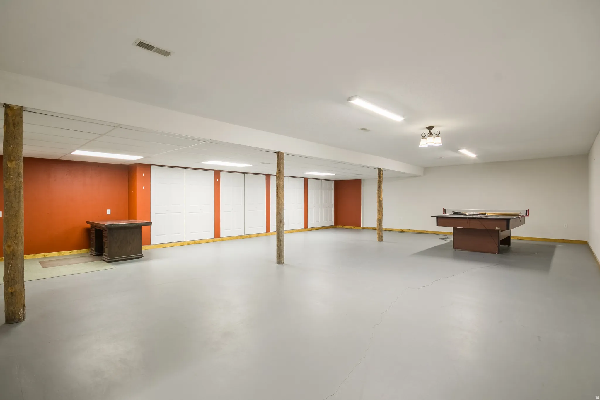 Spacious finished lower level featuring a light grey floor, drop ceiling with fluorescent lighting, and natural wood support columns