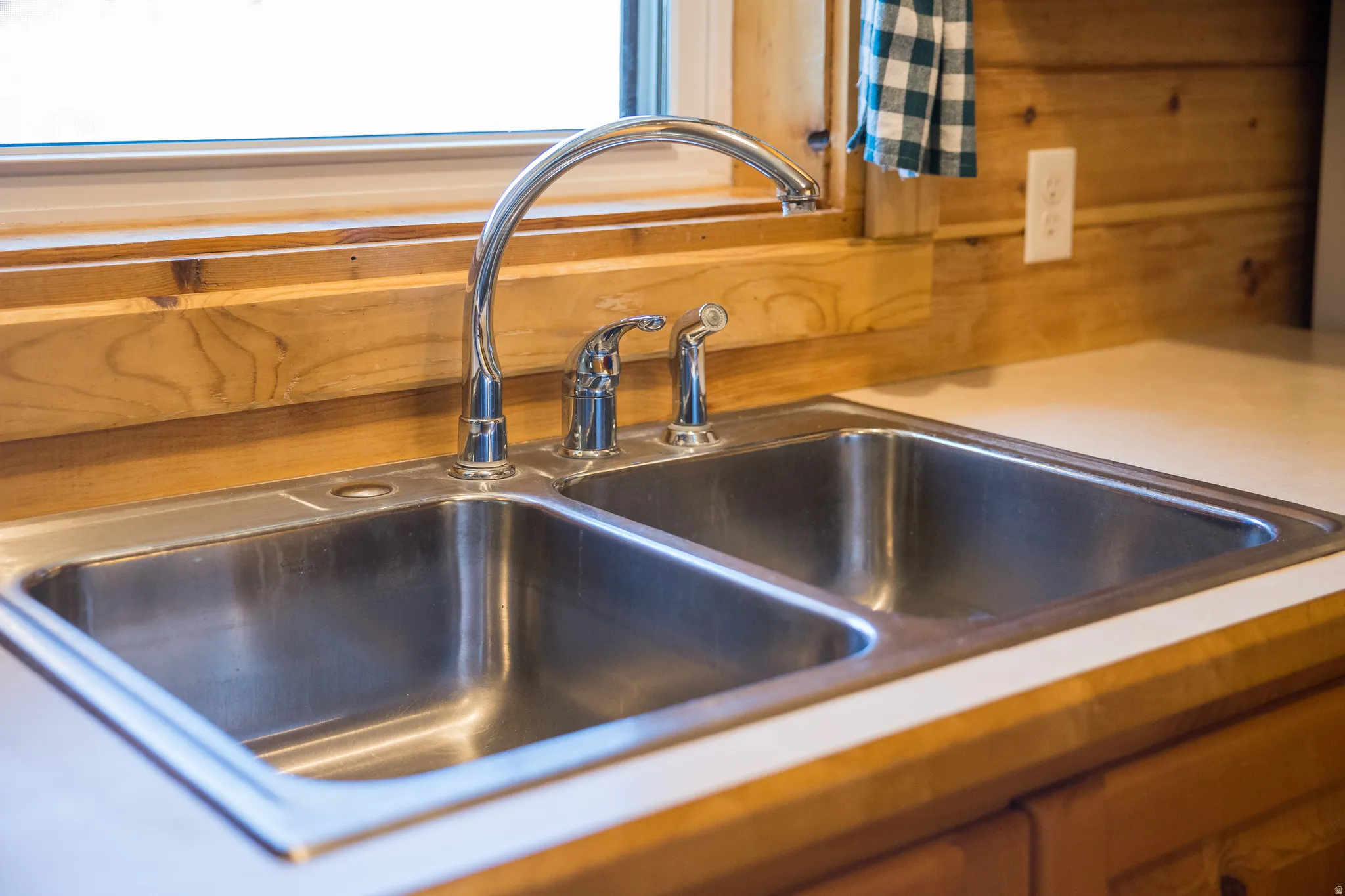 Double basin stainless steel sink with a high-arc chrome faucet, surrounded by light wood-finish cabinetry and a white countertop
