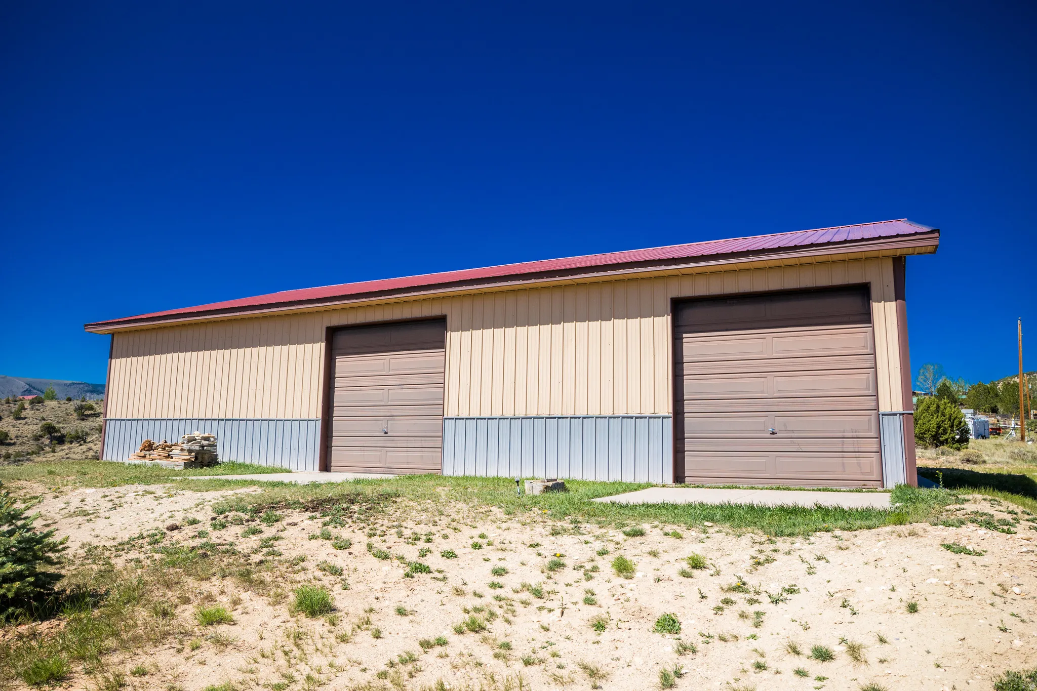 Metal building featuring a red-hued roof, two large garage doors, beige vertical siding, and a corrugated metal base