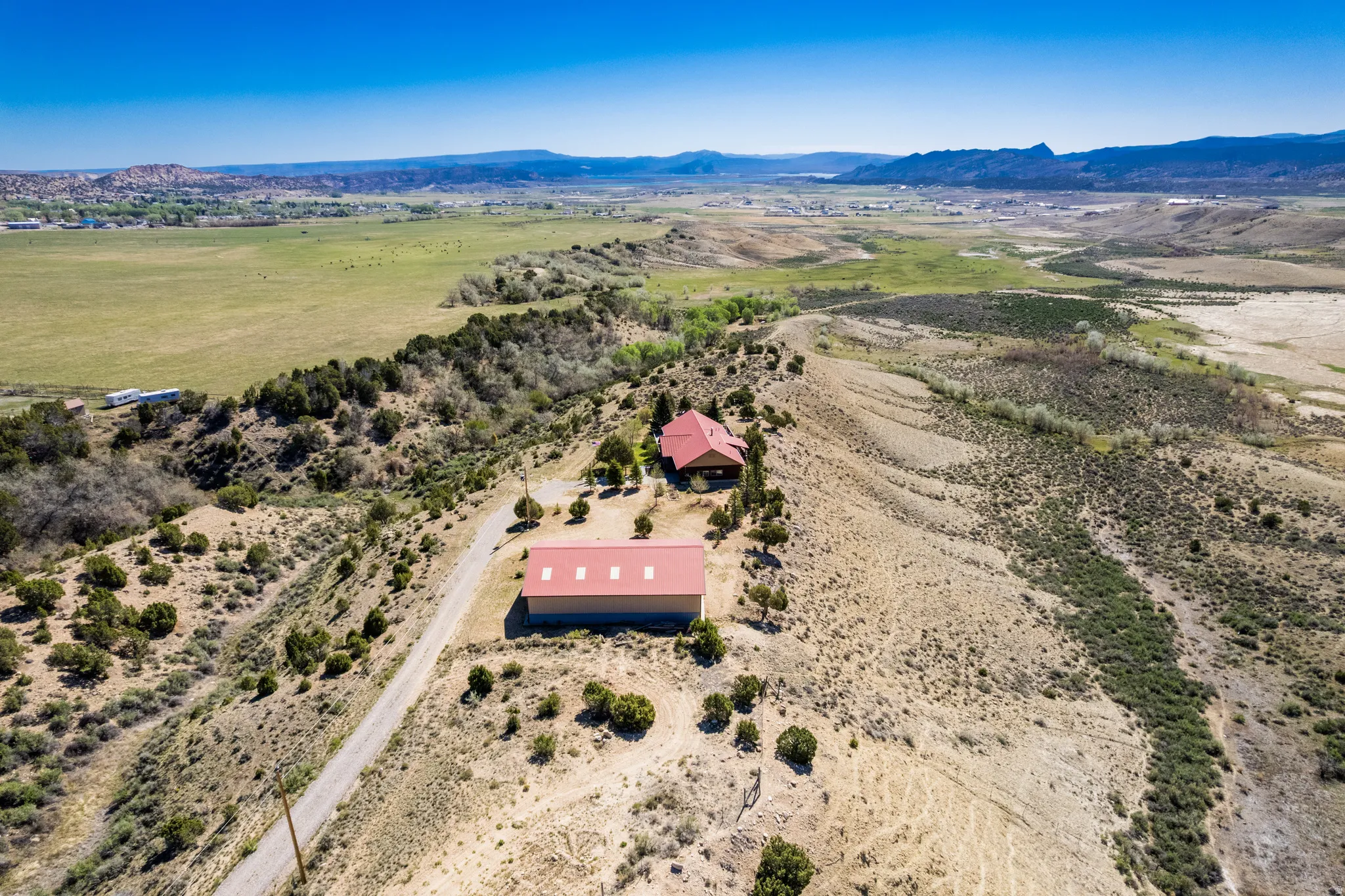 Expansive property with multiple structures featuring red metal roofs, a winding dirt road, and a mix of arid and green vegetation, all set against a distant mountainous backdrop