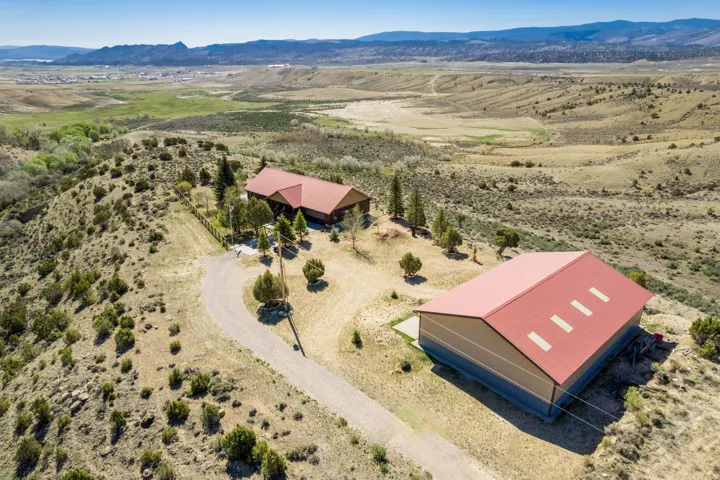 Aerial property view featuring a main residence with a red roof, a separate large outbuilding with a red roof, a gravel driveway, and surrounding natural landscaping on a hillside