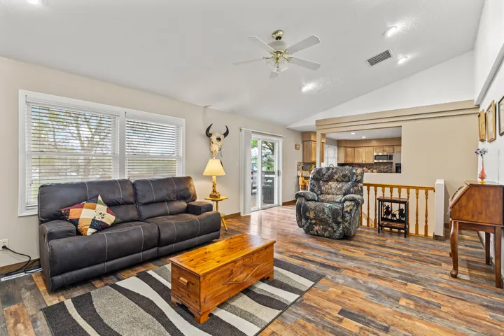 Vaulted ceiling great room with wood-finish flooring, two large windows with blinds, a ceiling fan, and recessed lighting