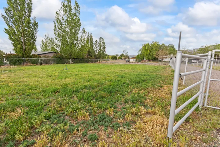 Expansive grassy lot featuring a metal gate, chain-link fencing, and mature trees lining the perimeter