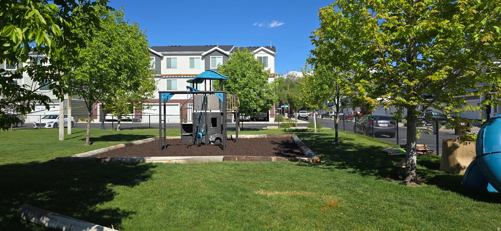 Community green space featuring a playground with a slide and climbing structure, surrounded by established trees, and backed by residential buildings