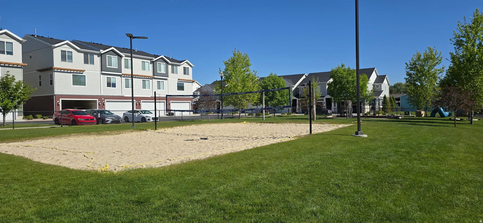 Expansive green space featuring a sand volleyball court, mature trees, and a playground area with a blue slide