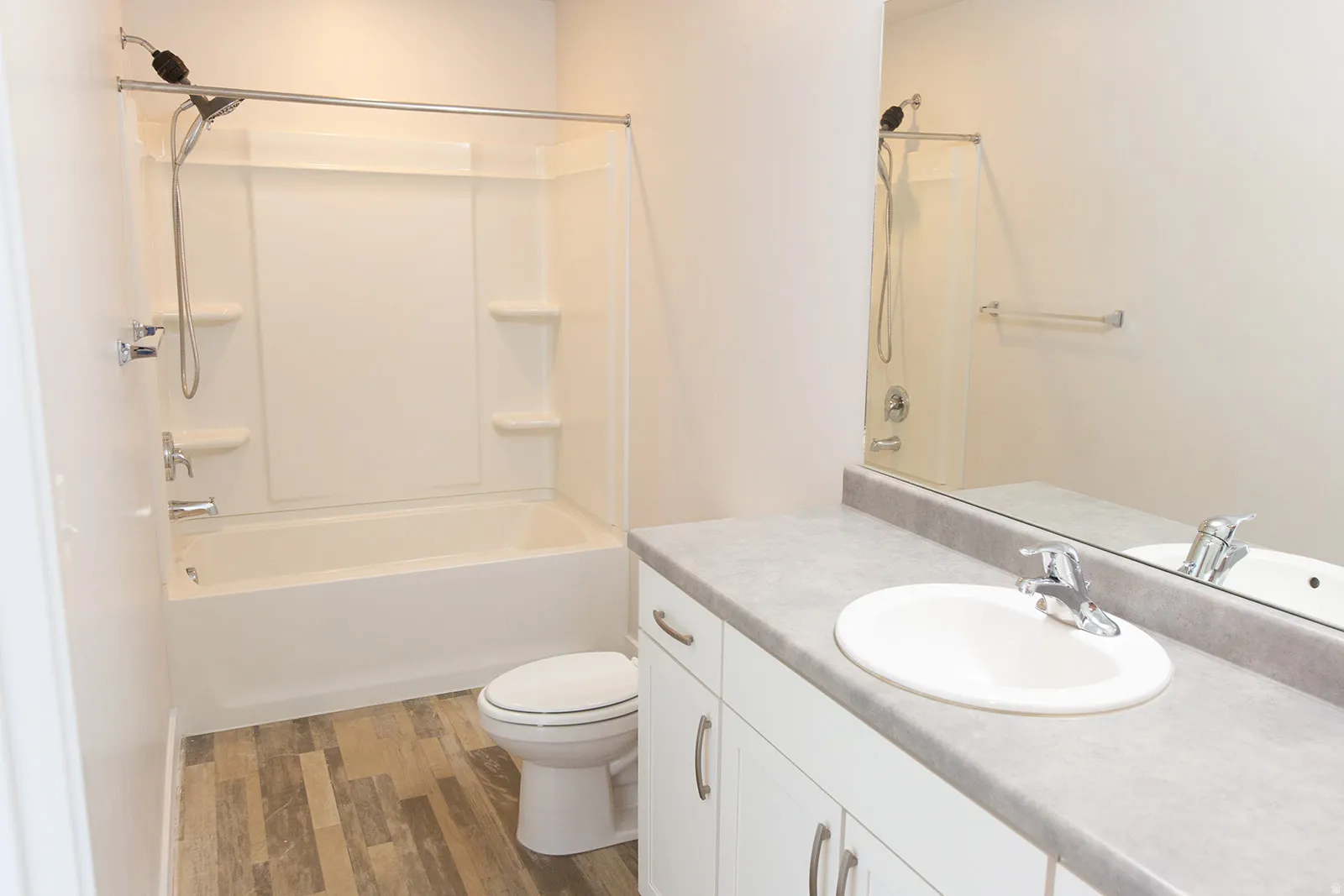 Bathroom featuring a white integrated tub and shower unit, a vanity with a white basin sink, a gray countertop, white cabinetry with silver pulls, and wood-finish flooring